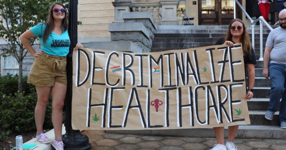 Two women hold a sign saying 'Decriminalize Healthcare'