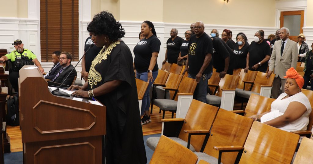 Residents of Pittard Road stand up in black shirts that say "Justice 31."